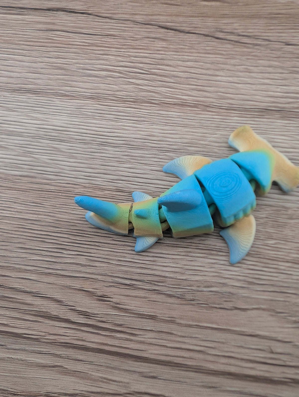 Articulated blue and beige hammerhead shark toy viewed diagonally, highlighting the wide head and segmented tail.