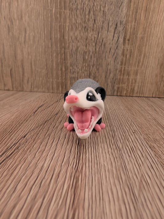 Gray articulated opossum figurine facing forward, mouth wide open in a defensive pose, resting on a wooden plank surface.