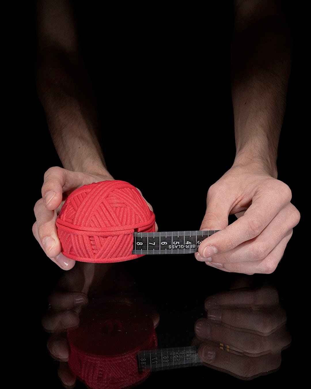 Two hands hold the matte red yarn ball object while measuring its diameter (about 7 cm) with a black fabric tape measure.