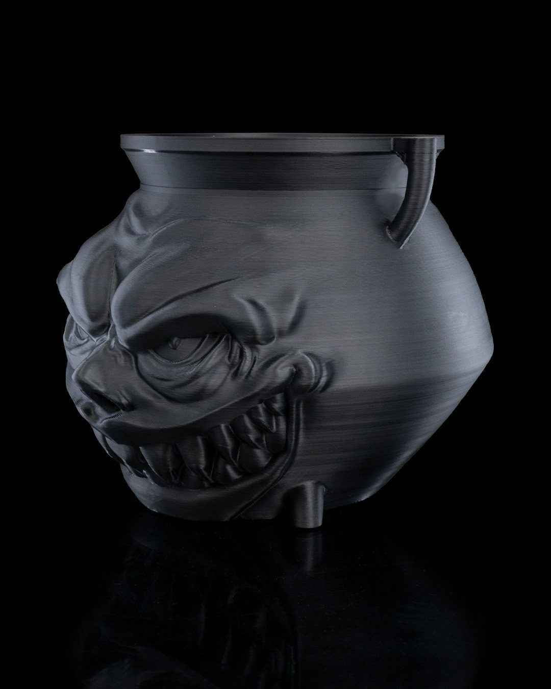 Side view of the dark metallic cauldron showing the extreme transition between the smooth body and textured monster face.