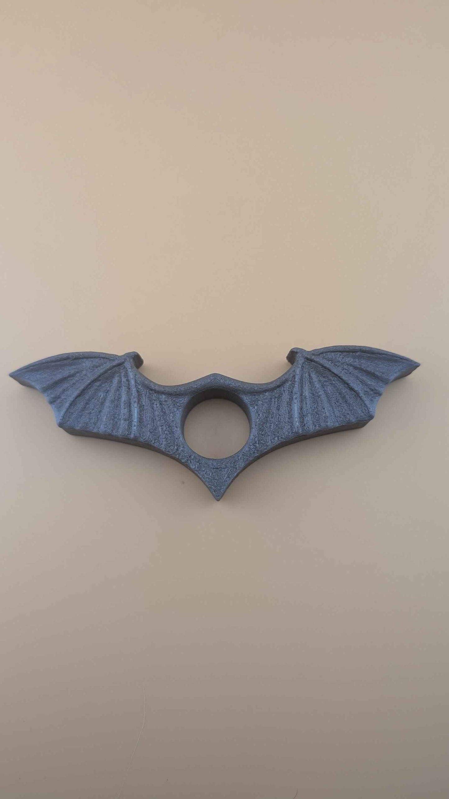 Top-down view of the symmetrical, matte dark grey bat wing thumb book clip against a plain tan background.