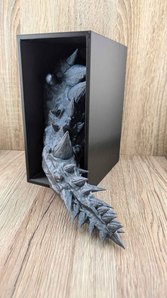Spiky dark grey metallic dragon tail sculpture emerging from a black box. Side view on wood grain surface.