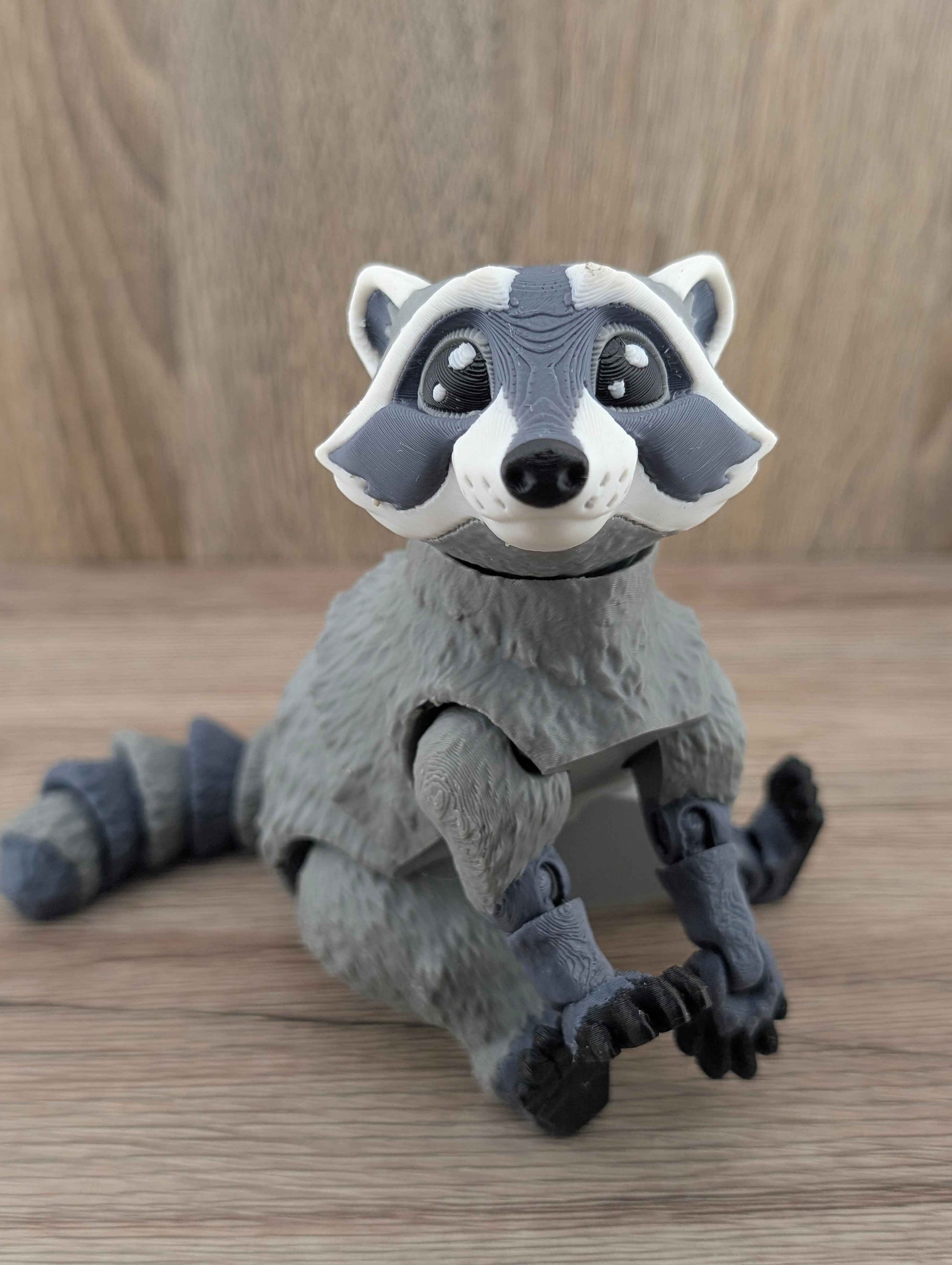 Gray raccoon figure sitting with its legs extended forward, showing the detailed textured fur body and striped segmented tail.