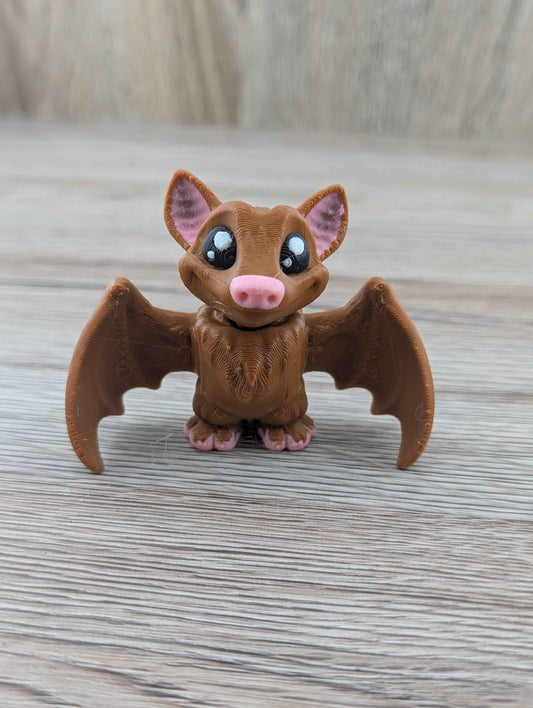Cute brown plastic bat figurine standing on a light wooden surface. Features a pink nose, pink ears, and large glossy eyes.