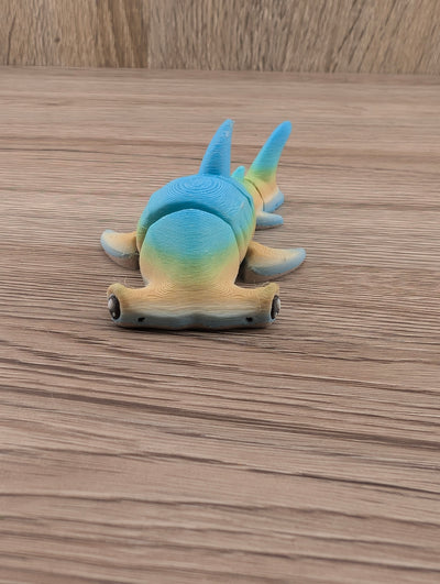 Articulated blue and yellow gradient hammerhead shark toy viewed from the front, resting on wood grain.