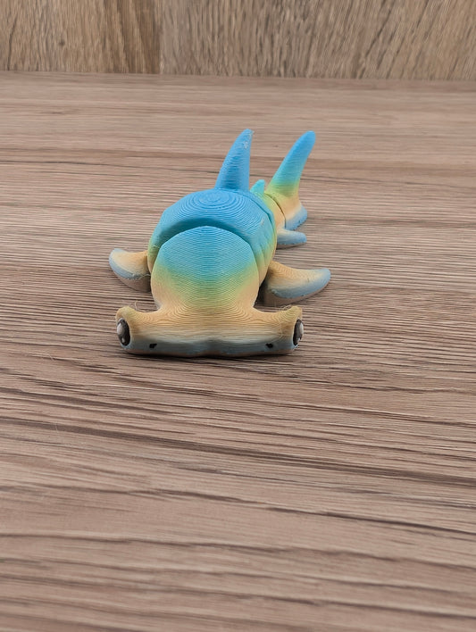 Articulated blue and yellow gradient hammerhead shark toy viewed from the front, resting on wood grain.