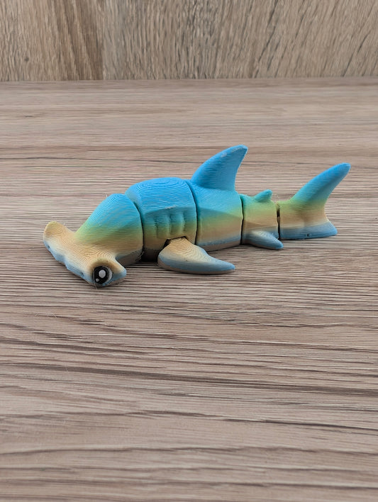 Side profile of a blue and tan segmented hammerhead shark. The body sections articulate, resting flat on a wooden surface.