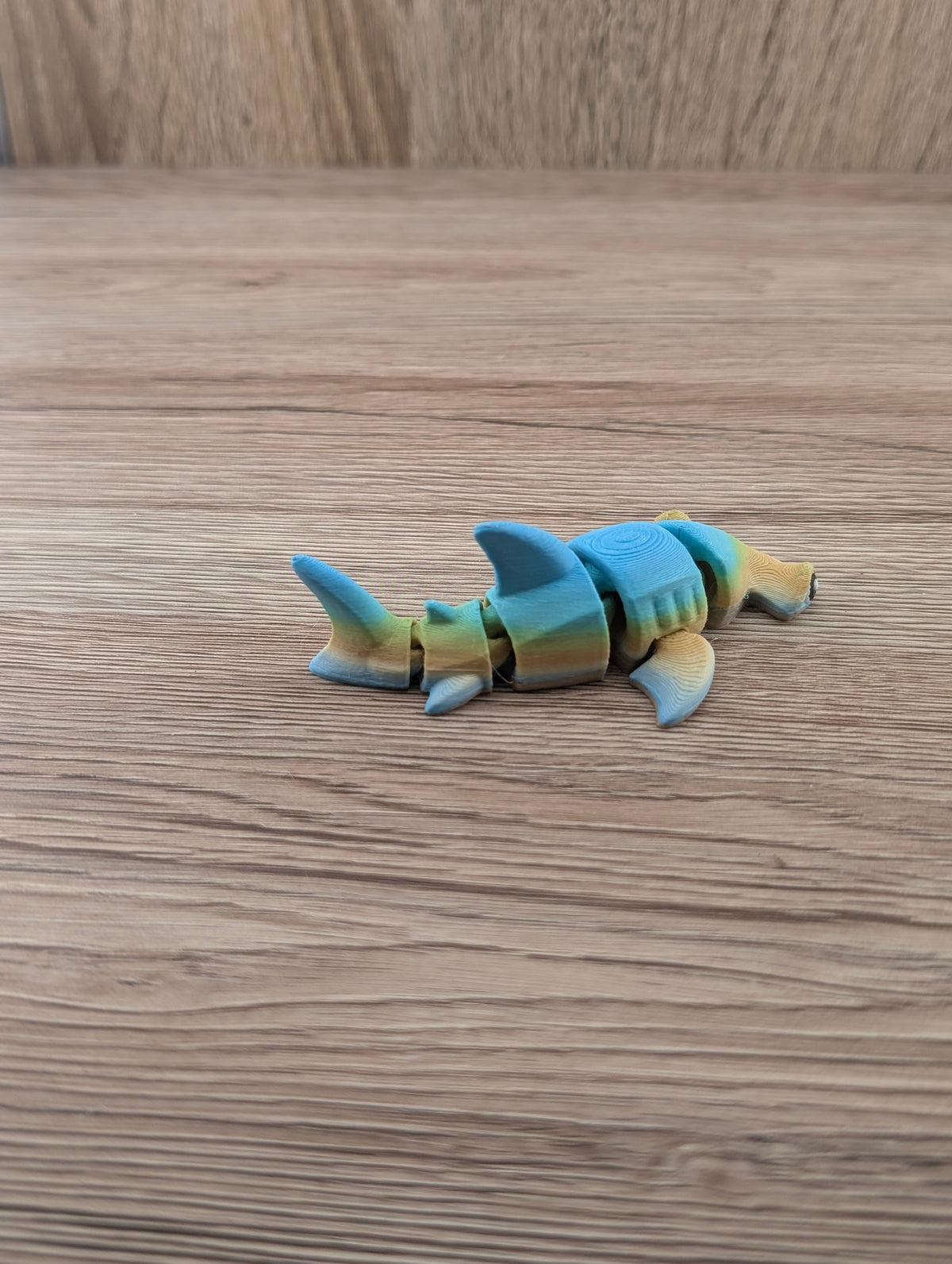 Segmented toy shark in blue, yellow, and tan colors, viewed from the side, showcasing layered texture on its fins.