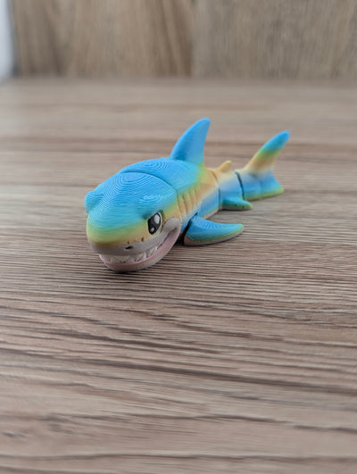 Cute articulated shark toy, multicolored blue, yellow, and beige, smiling with white teeth on a wood surface.