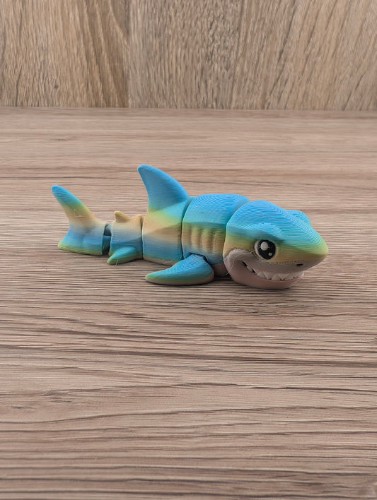 Side view of a flexible, segmented shark figure toy in blue and gold gradient colors on wood grain texture.