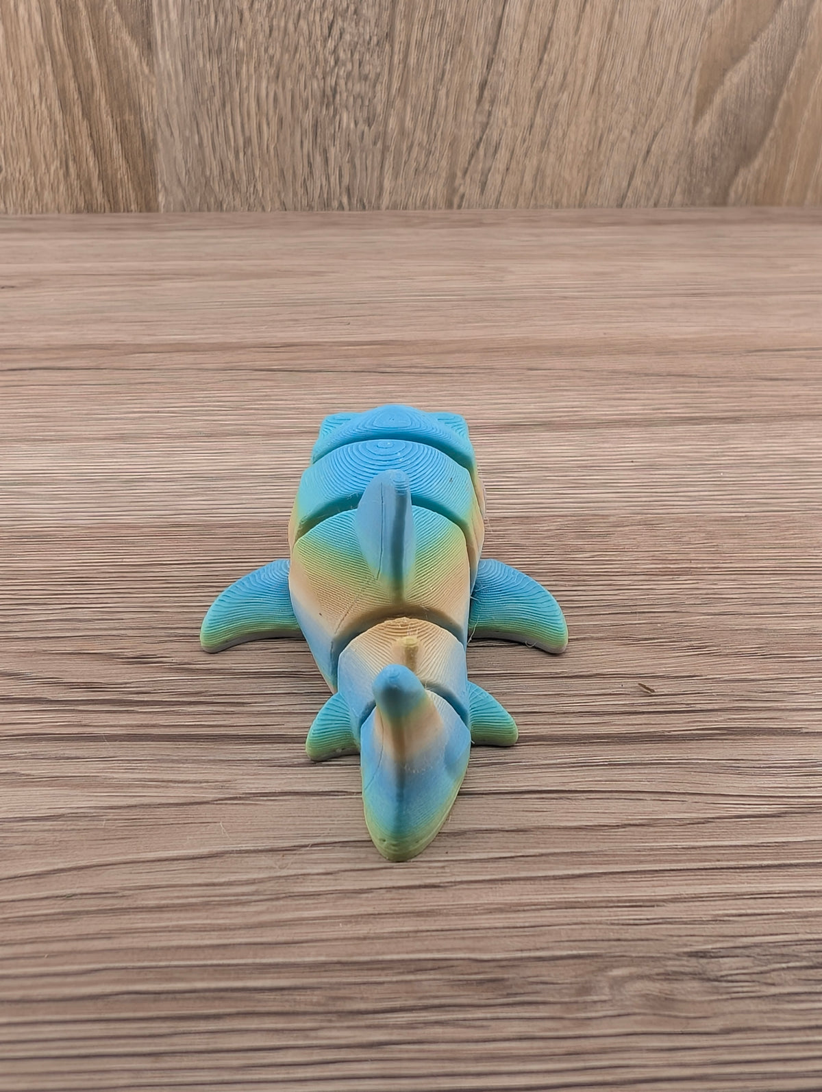 Head-on view of the ridged, multicolored shark toy with blue top and yellow underside resting on a wood table.