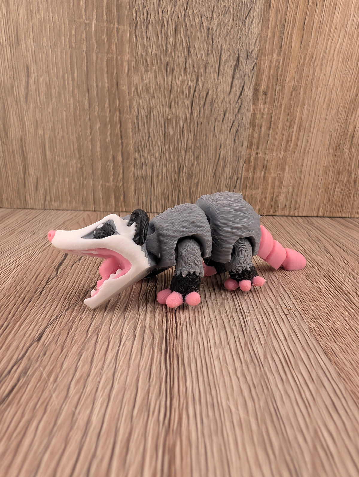 Profile view of an articulated gray opossum figurine with a textured body and an extended pink segmented tail, on wood.