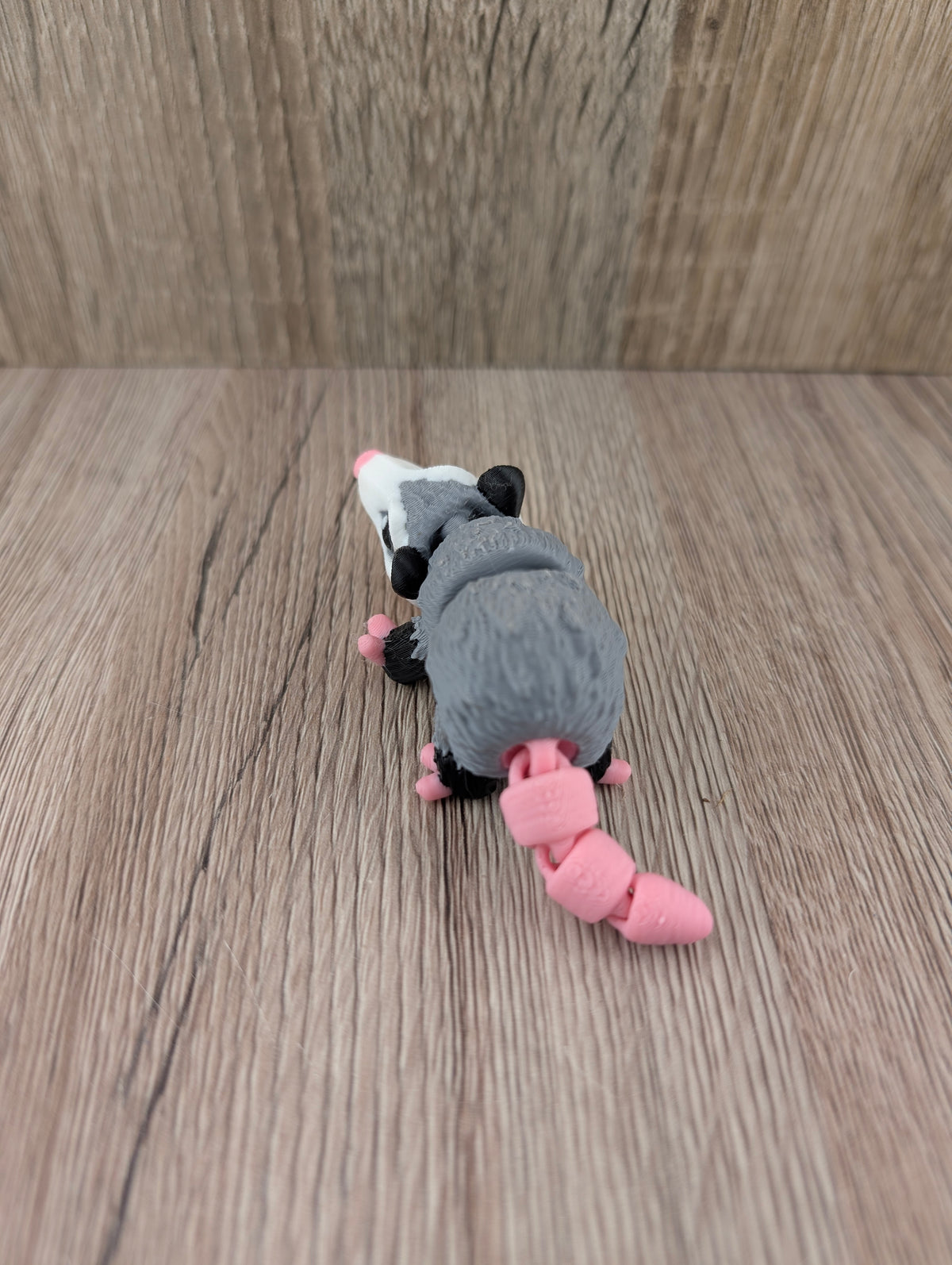 Overhead view of the articulated grey opossum toy, highlighting its textured segmented body and bright pink coiled tail.