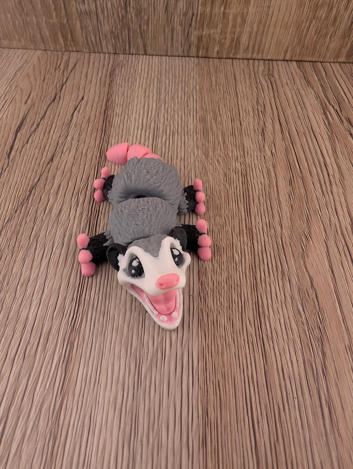 Articulated gray and white possum figurine lying on its back, simulating playing dead, with pink limbs spread wide.