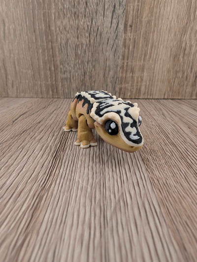 Articulated toy gecko with tan body and striking black and white striped shell segments, viewed on a wood surface.