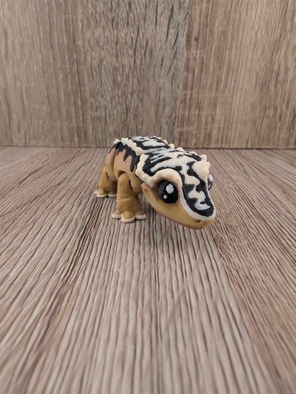 Articulated toy gecko with tan body and striking black and white striped shell segments, viewed on a wood surface.