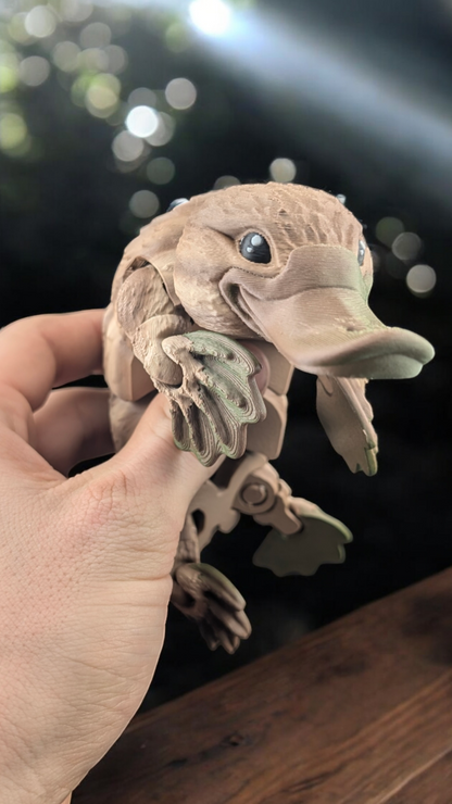 Close-up of a hinged platypus fidget toy held in a hand. It has a tan textured body and green webbed feet.