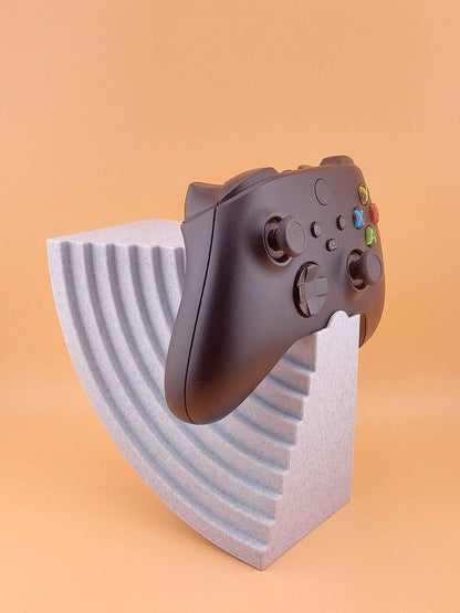 Black game controller resting on a speckled grey stand featuring a stepped, quarter-circle wave design, centred on a peach background.
