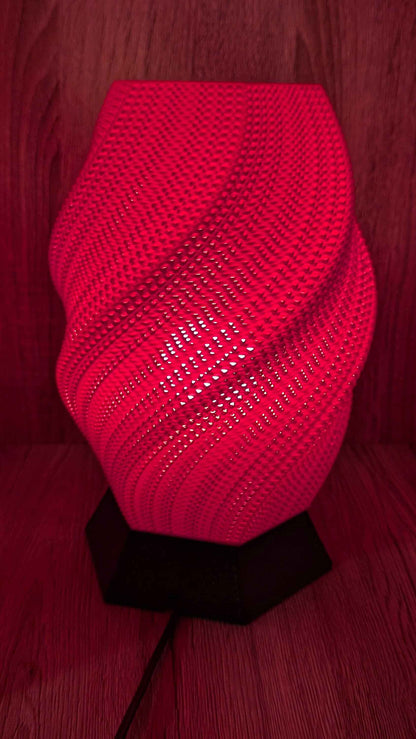 Twisted geometric lamp glowing intense red, revealing pinprick patterns where the light escapes the woven, dark red mesh.