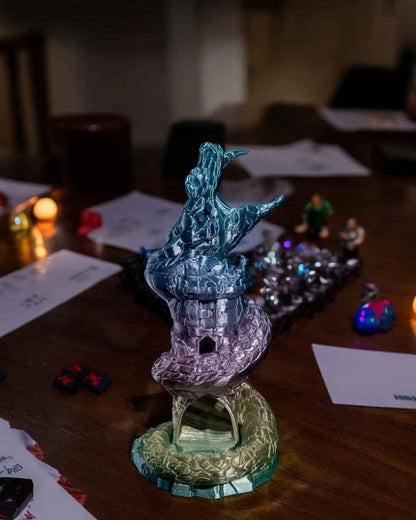 Iridescent serpentine tower dice roller viewed mid-shot on a dark table, shimmering teal down to gold in low light.