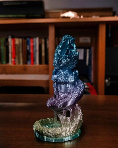 Serpentine tower dice roller in iridescent blue, purple, and gold, viewed straight on against a blurry bookshelf.