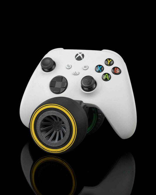 Studio shot, front view of a white Xbox controller resting on a matte black tire with a vibrant yellow rim.