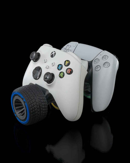 White Xbox and white PS5 controllers linked together, balanced on a single black treaded tyre with a blue rim.
