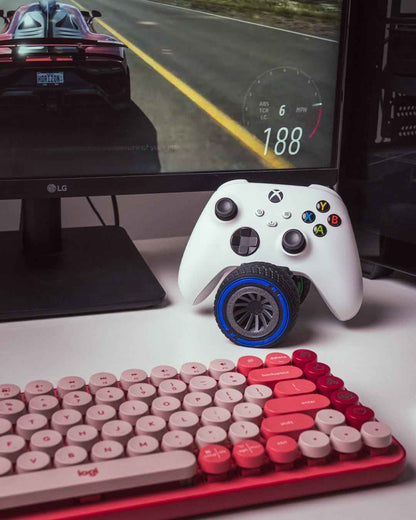 Action shot of a white Xbox controller on a blue-rimmed wheel, placed near a racing game screen and red keyboard.