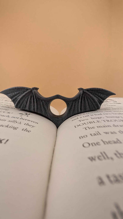 Dark charcoal grey bat wing book clip resting on the spine of an open book. Low angle showing the textured wing details.