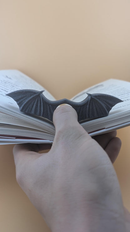 Hand holding the open book using the dark grey textured bat wing clip as a thumb page holder.