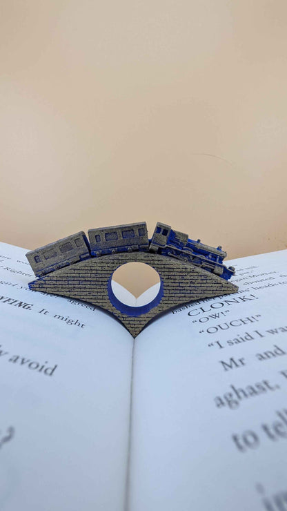 Detailed gold and blue train book holder crossing a textured brick bridge, inserted into the centre of an open book.