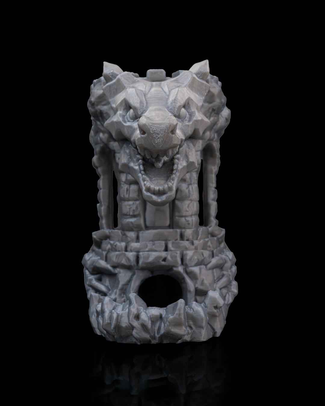Centred front view of the grey stone dice rolling tower. Features a snarling wolf or dragon head and a central exit hole below.