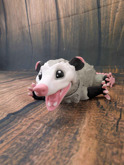 Flexible grey opossum figure laying flat on the ground with its limbs splayed, mouth wide open.