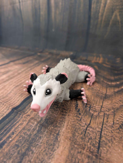 Full view of the grey opossum figure posed flat against the ground with limbs and pink tail spread out.
